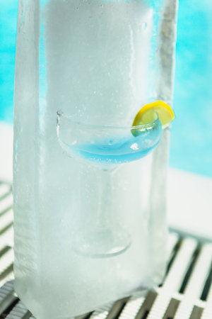 Blue cocktail in coupe glass with lemon garnish embedded in tall block of ice by pool
