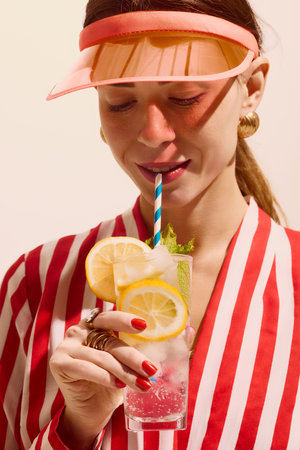Woman in striped shirt sipping citrus cocktail with visor and jewelryの写真素材