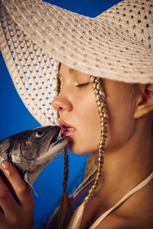 Close up woman in straw hat touching lips with fish against blue backgroundの写真素材