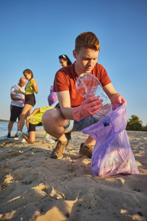 Young man in red shirt collecting plastic bottle on sandy beach with focus