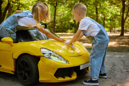 Children washing toy yellow car with sponge and foam in summer parkの写真素材