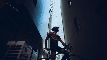 Cyclist standing with bike in narrow alleyway between tall modern buildings in dramatic lightの写真素材