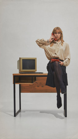 Confident woman in glasses holds red phone sitting at wooden desk with retro computer