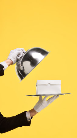 Silver cloche unveiling white copy space box on tray against yellow background. Mockup for ads