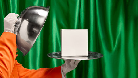Waiter hand lifting silver cloche to reveal white box on tray against green curtain backdrop