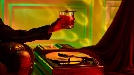 Elegant cocktail with cherry held in hand beside vintage record player in warm neon light