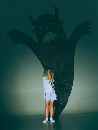 Creative digital artwork. Little girl covering eyes before giant devil shadow on green wallの写真素材