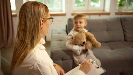 Psychologist observing boy with teddy bear during therapy session in soft daylightの写真素材