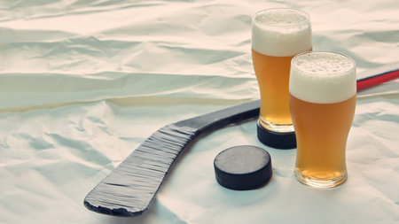 Two glasses of beer near hockey stick and puck on white backgroundの写真素材