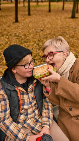 Grandmother sharing sandwich with grandson in autumn parkの写真素材
