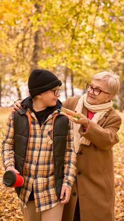 Grandmother walking with grandson in autumn park holding sandwich and coffeeの写真素材