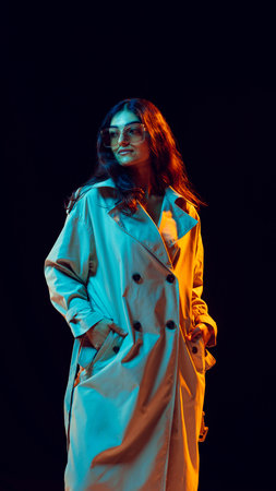 Woman in trench coat wearing sunglasses illuminated by blue and orange lightの写真素材