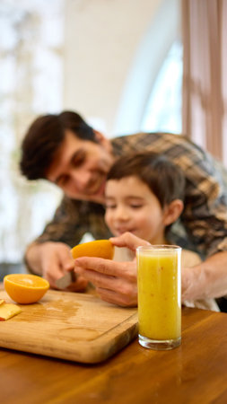 Fresh yellow smoothie in tall glass with father helping son cut orange in cozy kitchenの写真素材