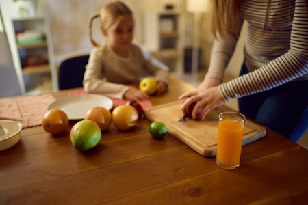 Mom cutting fresh fruits on wooden board with daughter and bright orange juice in cozy kitchen.の写真素材