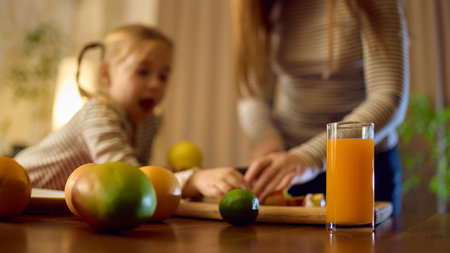 Bright orange juice standing on table during fruit preparation with joyful little girl and her momの写真素材