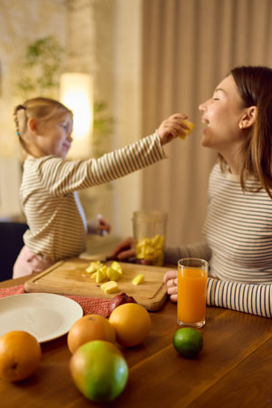 Orange juice on table while daughter feeds fruit to smiling mother in warm playful moodの写真素材