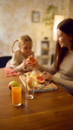 Orange juice glass on table while mother holds grapefruit halves with daughter nearbyの写真素材