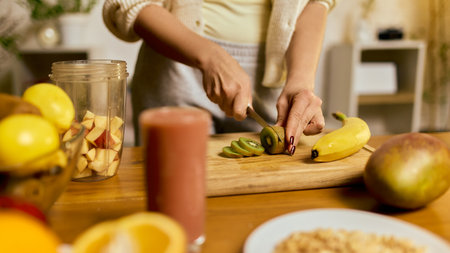Hands slicing ripe kiwi for smoothie preparation with fruits and blender on table