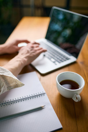 Hands typing on laptop beside notebook and cup of coffee on wooden table
