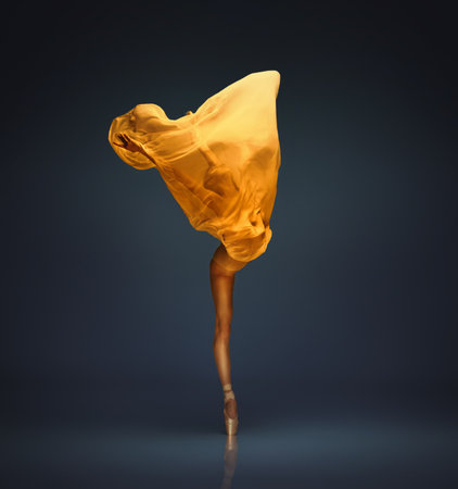 Ballet dancer lifting wide golden fabric shape on dark blue stage lightの写真素材