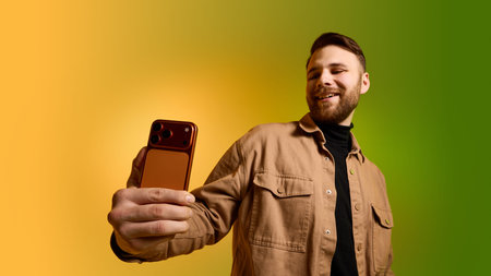 Bearded man taking selfie with smartphone on yellow green gradient background.