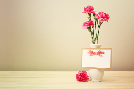 Pretty Pink Carnation Flowers on White Vase with Blank Greeting Card on Wooden Table with Light Brown Wall Backgroundの写真素材
