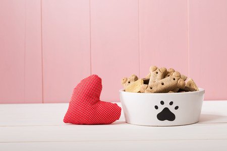 I Love My Dog concept with a red textile heart alongside a doggy bowl decorated with a paw print and filled with biscuits, pink background with copyspace
