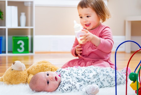 Happy toddler girl feeding her newborn baby sister a bottleの写真素材