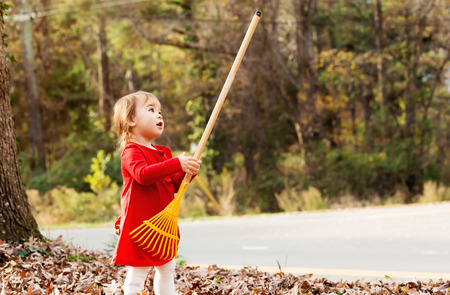 Toddler girl in a red shirt playing with a rake outside in autumnの写真素材
