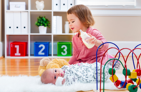 Happy toddler girl feeding her newborn baby sister a bottleの写真素材