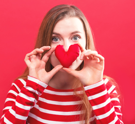 Happy young woman holding a heart cushion on a red backgroundの写真素材