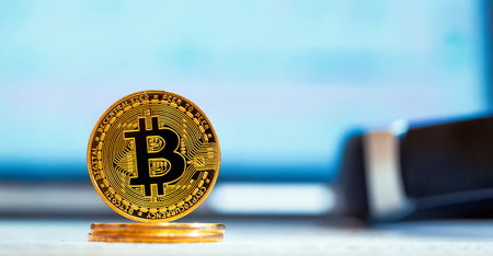 A gold bitcoin cryptocurrency coin with a laptop screen in the background