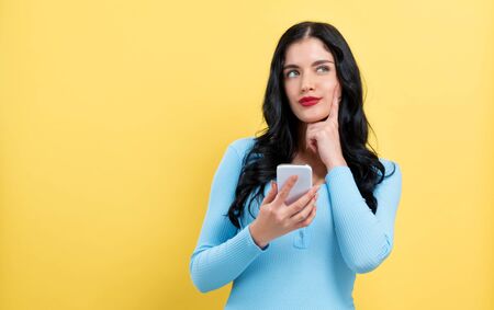 Happy young woman with smart phone thinking about something on a yellow backgroundの写真素材
