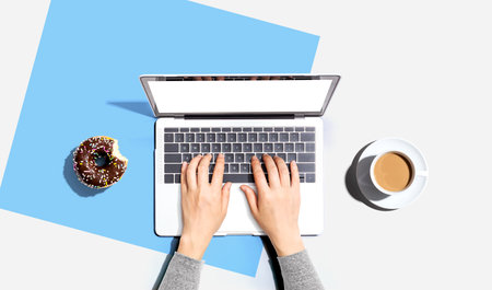 Person using a laptop computer with a donut and a cup of coffeeの写真素材