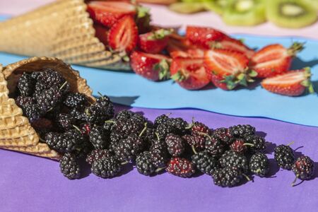 Scattered fruits (strawberries, pineapple, raspberries, kiwi, blackberries, cherries) in waffle, cone cups on a bright colored backgroundの写真素材