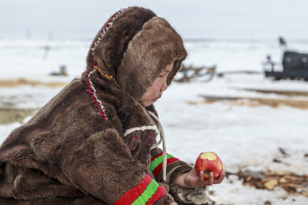 Nadym, Russia - april 27, 2018:tundra, open area, the boy is eating apple in cold winter weather, the boy  in national clothes, editorial.のeditorial素材