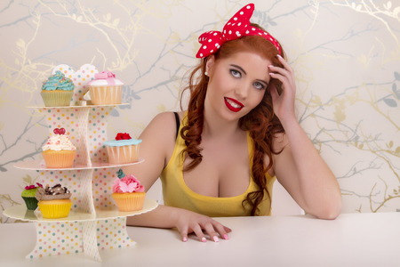 View of a beautiful pinup redhead girl with colorful cupcakes on a table.の写真素材