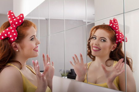 View of a beautiful redhead girl with colorful pinup clothes next to a mirror wall.の写真素材
