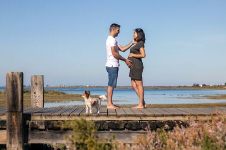 Pregnant happy couple with small dog poses near the marshlands.
