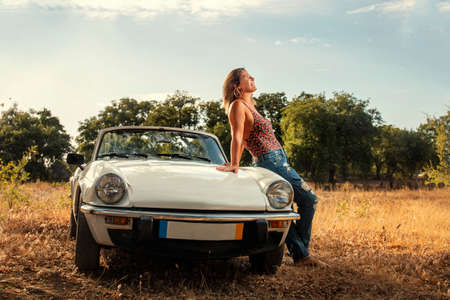 Woman with white convertible vintage car on the countryside, in posing fashionable lifestyle.