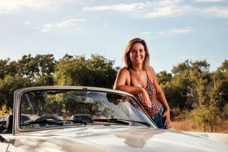 Woman with white convertible vintage car on the countryside, in posing fashionable lifestyle.