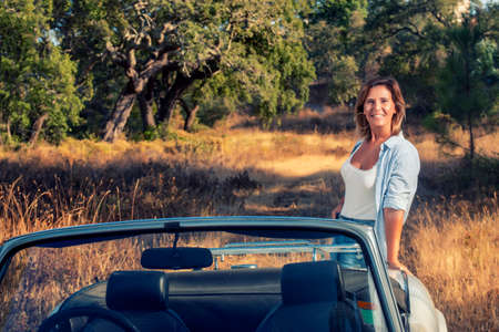 Woman with white convertible vintage car on the countryside, in posing fashionable lifestyle.