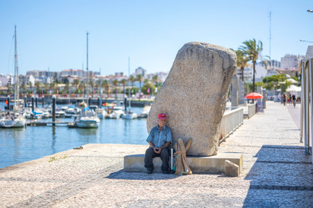 PORTIMAO, PORTUGAL - 29th JULY 2023: View of Docks with several anchored boats in the city of Portimao, Algarve, Portugal. Old man relaxes next to big stone artwork.のeditorial素材