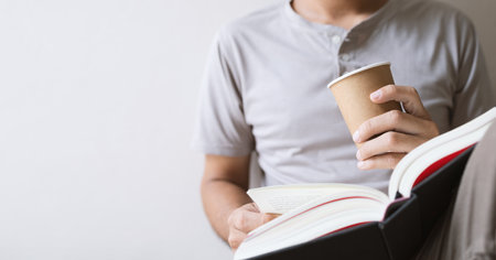 young man is sitting reading a book while holding a paper cup of coffee in the room with soft-focus and over light in the backgroundの写真素材