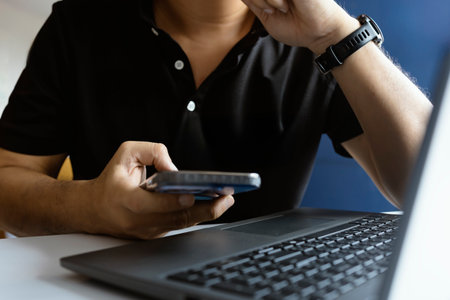 Closeup of hand using smartphone with laptop on desk, illustrating digital communication, mobile technology, and modern workflow.の写真素材