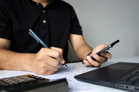 Man filling out tax form using pen and phone, with calculator and laptop on white desk
