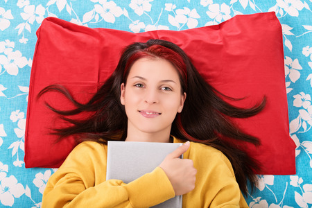 A portrait shot of a beautiful young girl lying in her bed and holding a book.の写真素材