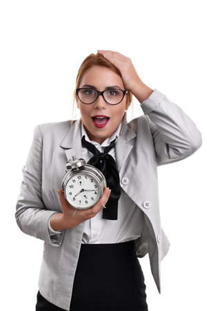 Beautiful young business woman dressed in a suit holding a clock with shocked expression, isolated on a white background.の写真素材