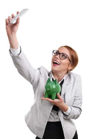 Smiling beautiful young business woman dressed in a suit with glasses taking a selfie with a green piggy bank, isolated on a white background.の写真素材