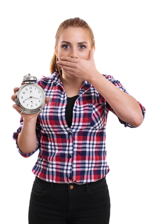 Portrait of a young beautiful woman dressed in plaid shirt holding an old school alarm clock, shocked and surprised to see what's the time. Isolated on a white background.の写真素材
